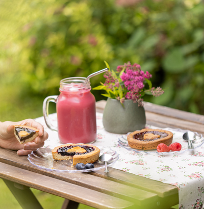 Une table de jardin avec un goûter composé d'un grand verrre de délicieux Smoothie Framboise et de deux Tartelettes façon Linzer, Tartelette Framboise et Tartelette Myrtille.